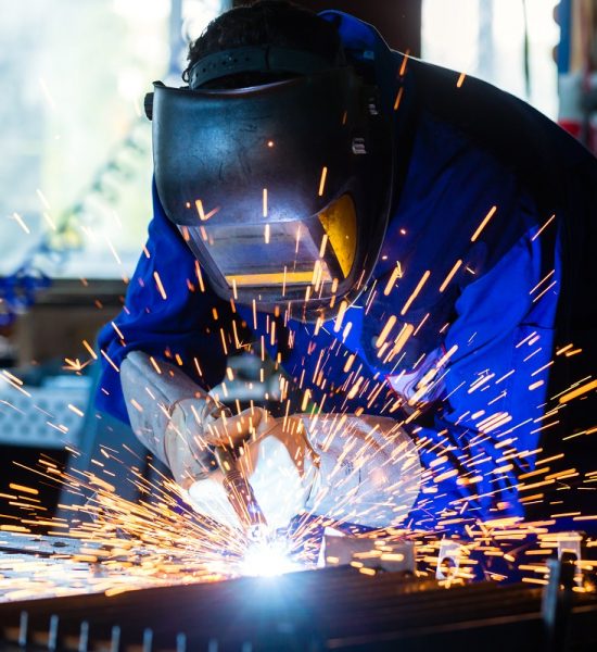 Welder bonding metal with welding device in workshop, lots of sparks to be seen, he wears welding googles